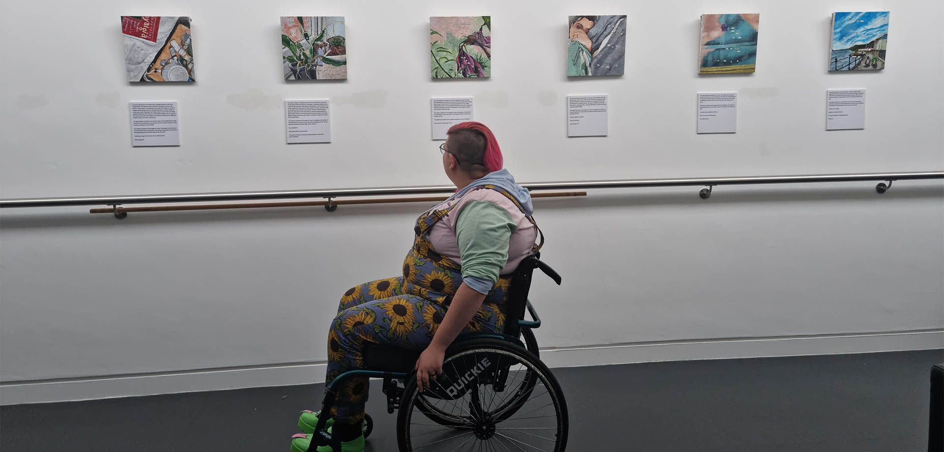 A person in a wheelchair in the foreground is turning away to look at six square paintings hung on a white wall.
