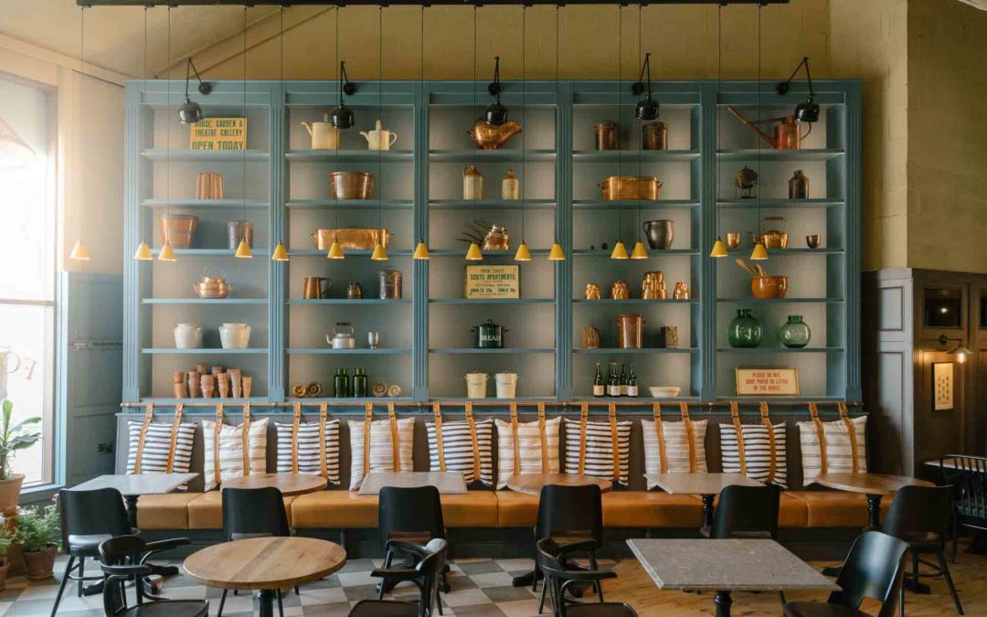 Interior of Chatsworth Kitchen showing blue shelves piled with jelly moulds, old signage and other Chatsworth historical artefacts, banquette seating with striped cushions, wooden tables and metal chairs, sun streams through the window