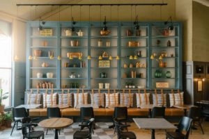 Interior of Chatsworth Kitchen showing blue shelves piled with jelly moulds, old signage and other Chatsworth historical artefacts, banquette seating with striped cushions, wooden tables and metal chairs, sun streams through the window