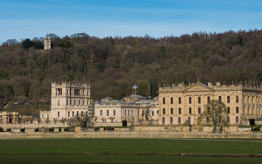 Chatsworth House in the foreground with a dense woodland behind it and the hunting tower visible