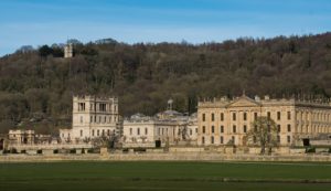Chatsworth House in the foreground with a dense woodland behind it and the hunting tower visible